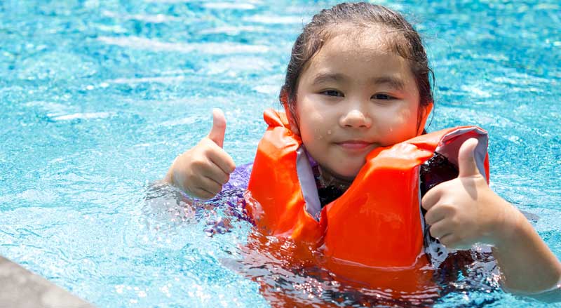 a young child in a life jacket is learning water safety and gives two thumbs up.